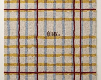 May include: A rectangular area rug featuring a cream-colored base with a grid pattern of intersecting lines. The design includes vertical and horizontal stripes in light blue and mustard yellow, overlaid with bold, dark red grid lines. STR RUGS logo.