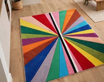 May include: A rectangular area rug featuring a vibrant sunburst pattern composed of numerous triangular segments in a wide spectrum of colors including red, orange, yellow, green, blue, purple, and pink, separated by a central black and white stripe.