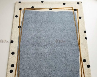 May include: A rectangular area rug featuring a cream-colored background with scattered black polka dots. The center displays a large, textured, light blue rectangular shape outlined by two overlapping thin, dark gold-colored rectangular frames.