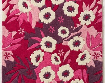 May include: A rectangular rug featuring a dense floral pattern in shades of magenta, deep purple, light pink, and cream. The design consists of stylized flowers with dark purple centers and overlapping leaves, creating a textured, layered appearance.