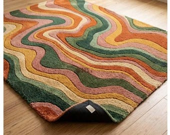 May include: A rectangular area rug featuring a wavy, multi-colored pattern of orange, forest green, mustard yellow, and dusty pink. The rug has a textured, tufted surface and one corner is folded back to reveal a black backing with a white label.