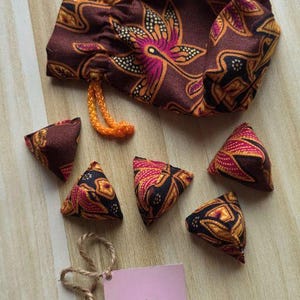 May include: A brown fabric drawstring pouch with an orange cord and five matching triangular fabric bean bags featuring a floral pattern in pink, orange, and yellow. A square pink tag with a black smiley face and the text TheLittleStitchSmile.