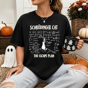 Puede incluir: Una camiseta negra de manga corta con texto blanco que dice SCHRÖDINGER CAT y THE ESCAPE PLAN, con un gráfico central de la silueta de un gato rodeado de complejas ecuaciones de física y la ilustración de un pequeño cohete.