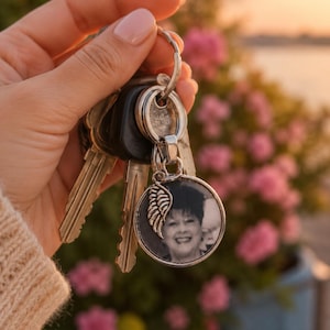 May include: A silver keychain with a round pendant featuring a black and white photo of a smiling person. A silver wing charm is attached to the pendant. The keychain holds several keys.