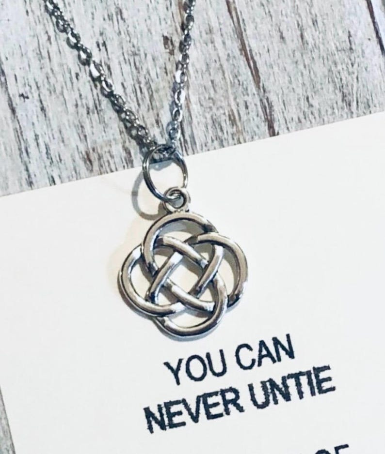 Puede incluir: Un collar plateado con un colgante de nudo celta. El colgante tiene un dise&ntilde;o de tr&eacute;bol de cuatro hojas. El collar est&aacute; sobre una tarjeta blanca con el texto "YOU CAN NEVER UNTIE".