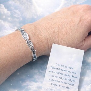 Puede incluir: Pulsera plateada con alas de &aacute;ngel. La pulsera se lleva en la mu&ntilde;eca, con una tarjeta que dice "You left me with beautiful memories. Your love is still my guide..." sobre un fondo de cielo nublado.