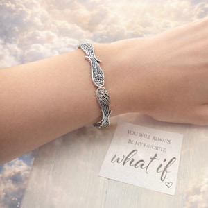 May include: A silver-toned bracelet featuring a wing design. The bracelet is worn on a wrist. A small card with the text "YOU WILL ALWAYS BE MY FAVORITE what if" is visible.