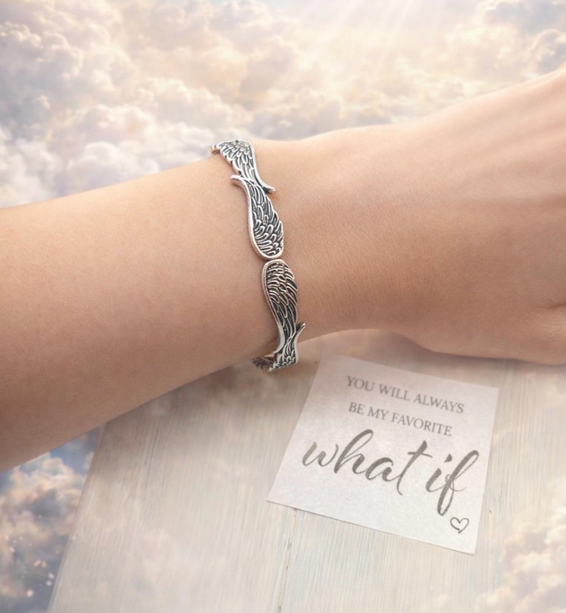 May include: A silver-toned bracelet featuring a wing design. The bracelet is worn on a wrist. A small card with the text "YOU WILL ALWAYS BE MY FAVORITE what if" is visible.