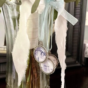 May include: Two silver photo charms with black and white photos of a couple and an older person. The charms are hanging from a bouquet of flowers in a clear glass vase with a white and blue ribbon.
