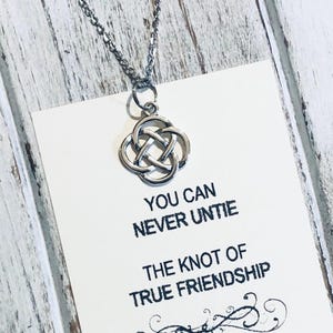 Puede incluir: Un collar plateado con un colgante de nudo celta cuelga sobre una tarjeta blanca. La tarjeta tiene el texto "YOU CAN NEVER UNTIE THE KNOT OF TRUE FRIENDSHIP" en negro. El fondo es de madera clara y desgastada.