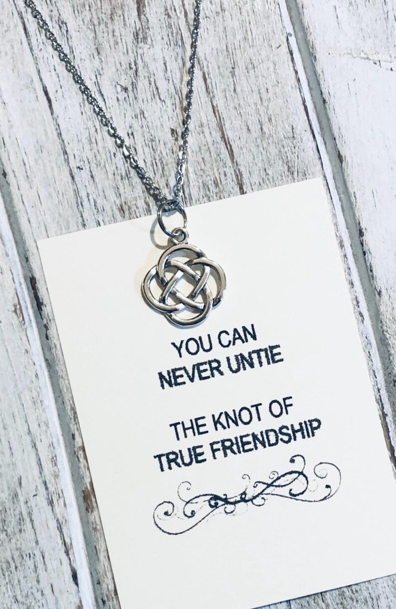 Puede incluir: Un collar plateado con un colgante de nudo celta cuelga sobre una tarjeta blanca. La tarjeta tiene el texto "YOU CAN NEVER UNTIE THE KNOT OF TRUE FRIENDSHIP" en negro. El fondo es de madera clara y desgastada.