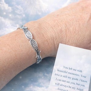 Puede incluir: Una pulsera plateada con dise&ntilde;o de alas se lleva en la mu&ntilde;eca. La pulsera presenta patrones detallados de plumas. Una tarjeta con texto es visible, sobre un fondo de cielo nublado.