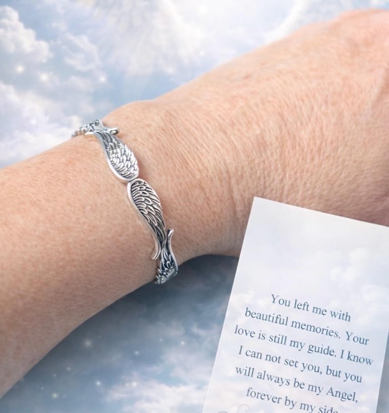 Puede incluir: Una pulsera plateada con dise&ntilde;o de alas se lleva en la mu&ntilde;eca. La pulsera presenta patrones detallados de plumas. Una tarjeta con texto es visible, sobre un fondo de cielo nublado.