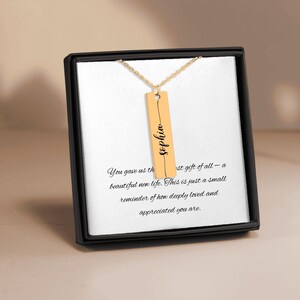 May include: A gold-coloured necklace with a rectangular pendant engraved with the name "Sophia" in cursive. The necklace is presented in a black gift box with a white card that has text on it.