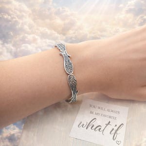 May include: Silver-toned bracelet featuring a wing design. The bracelet is worn on a wrist, with a card that reads "YOU WILL ALWAYS BE MY FAVORITE what if". The background shows a cloudy sky.