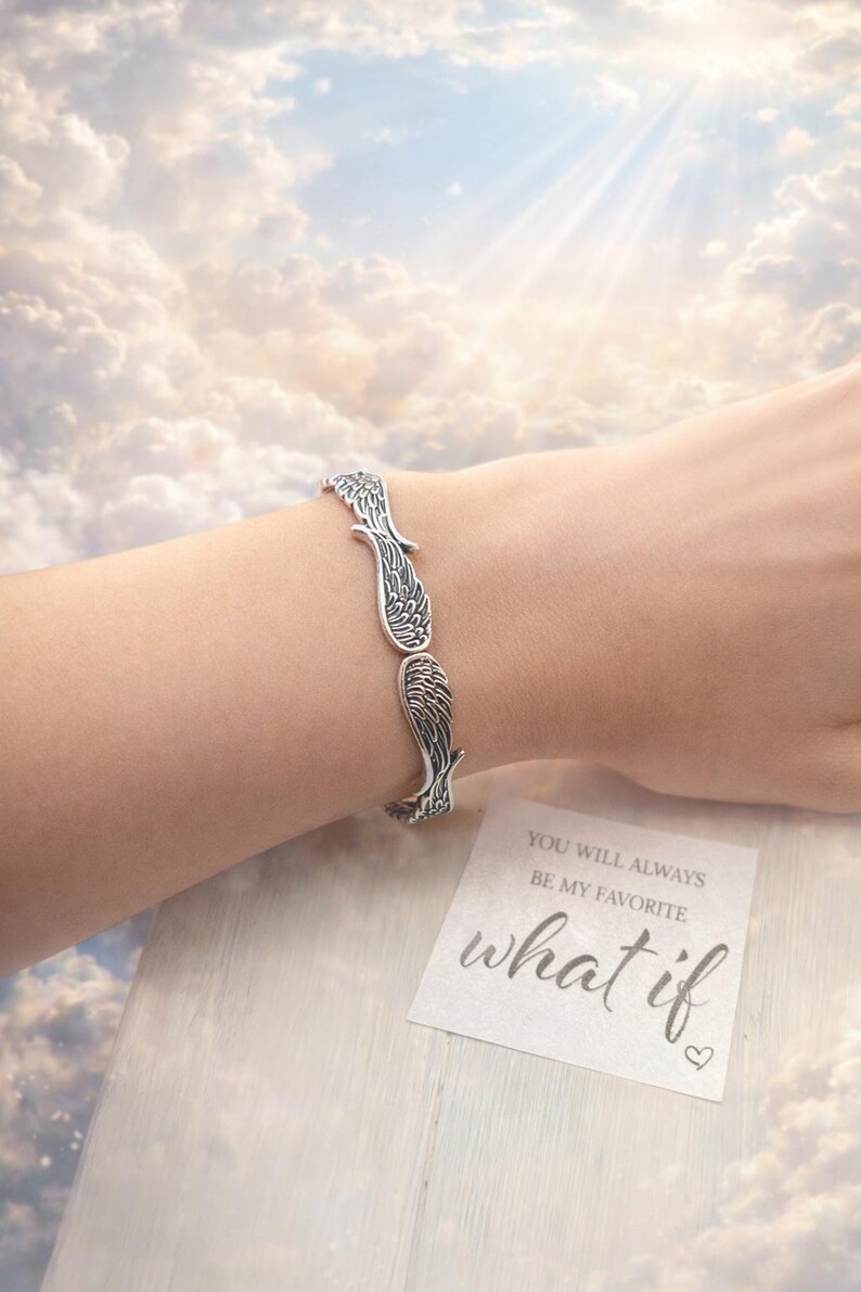 May include: Silver-toned bracelet featuring a wing design. The bracelet is worn on a wrist, with a card that reads "YOU WILL ALWAYS BE MY FAVORITE what if". The background shows a cloudy sky.