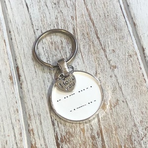 May include: A silver keychain with a round white charm featuring the words "I love you" in braille. The charm is attached to a silver heart charm with the words "I love you" engraved on it.
