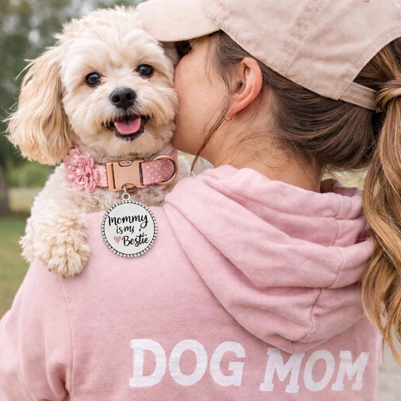May include: A cream-coloured dog with a pink collar and a tag that reads "Mommy is my Bestie." The dog is being held by a person wearing a pink hoodie with "DOG MOM" printed on the back. The dog has a fluffy coat.