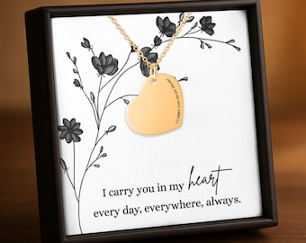 Può includere: Una collana con ciondolo a forma di cuore dorato su una catenina sottile, presentata in una scatola nera con un cartoncino bianco con illustrazioni floreali nere e il testo: I carry you in my heart every day, everywhere, always.