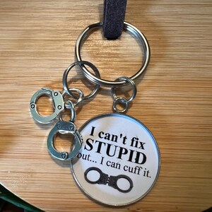 May include: Keychain with a round pendant and miniature handcuffs. The pendant has the text "I can't fix STUPID out... I can cuff it." The keychain has a silver-colored ring and a brown leather strap.