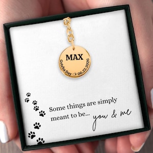 May include: A gold-toned circular pendant necklace featuring the name MAX, the text Gotcha Day, and the date 06.22.2026 with a small paw print icon. The pendant is presented in a black box with a card printed with paw prints and the text Some things are simply meant to be... you & me.