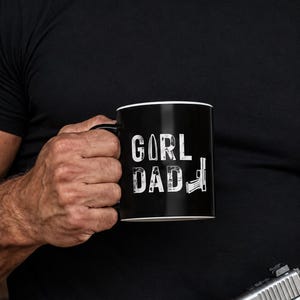 May include: A black ceramic mug featuring white distressed text that reads GIRL DAD. The letter I in GIRL is replaced by a bullet graphic, and the letter D in DAD is replaced by a handgun graphic. The mug has a white interior rim.