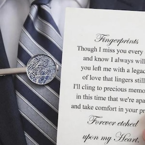 May include: A silver tie clip featuring a circular inlay with a dark blue fingerprint pattern, worn on a navy and silver striped necktie. Beside it is a white card with black text that reads: Fingerprints, Though I miss you every day and know I always will, you left me with a legacy of love that lingers still. I'll cling to precious memories in this time that we're apart, and take comfort in your presence, Forever etched upon my Heart.