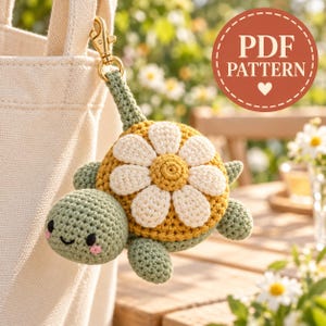 May include: A crochet turtle keyring featuring a light green head with pink cheeks and a shell designed as a yellow and white daisy flower. The turtle is attached to a gold-toned metal lobster clasp. The image includes the text PDF PATTERN.