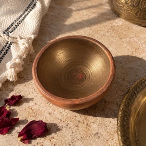 May include: A small, round bowl with a smooth, metallic gold interior and a matte, terracotta-colored exterior. The bowl features a circular rim and a deep, concave basin. A small, faint red mark is visible on the bottom interior surface.