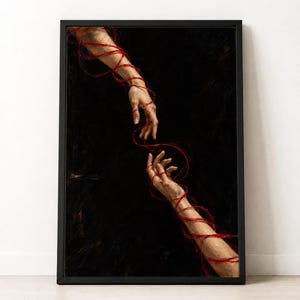 Op de afbeelding: Een ingelijste muurkunstprint met een schilderij van twee handen die naar elkaar reiken tegen een donkere zwarte achtergrond. Beide armen en handen zijn omwikkeld met een dunne, felrode draad die tussen de vingers doorloopt en verbindt.