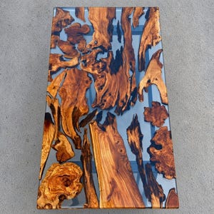 May include: A rectangular coffee table featuring a live edge wood slab design with natural brown wood grain patterns embedded in a clear, transparent resin surface. The wood pieces are arranged in an organic, irregular layout across the top.