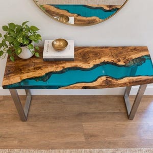 Peut inclure: Une console rectangulaire composée d'un plateau en bois brut à bords naturels avec une incrustation centrale en résine turquoise translucide. La table repose sur deux pieds en métal brossé en forme de U et porte une plante et un livre.