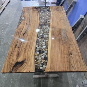 May include: A rectangular dining table featuring two natural live-edge wood slabs with a prominent grain pattern. A central channel filled with smooth, multi-colored river stones is encased in a clear, glossy resin finish.
