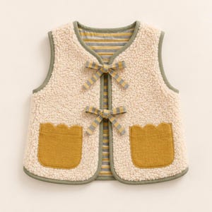 Può includere: Un gilet senza maniche in pile sherpa color crema con due tasche frontali applicate in giallo senape con bordi smerlati. Il gilet presenta una fodera a righe grigie e gialle e due lacci in tessuto a righe abbinati sul davanti.