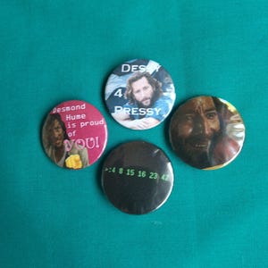 Puede incluir: Cuatro chapas redondas con imágenes de la serie de televisión Lost. Una chapa es rosa con el texto Desmond Hume is proud of you. Otra muestra a un hombre con el texto DES 4 PRESSY. Una presenta el rostro de un hombre y otra es negra con los números verdes 4 8 15 16 23 42.