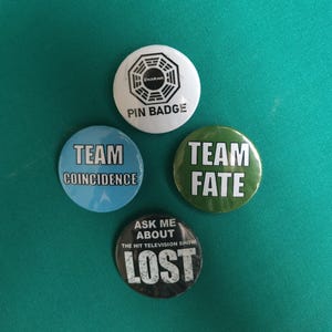 Könnte beinhalten: Vier runde Anstecknadeln. Eine weiße mit schwarzem Dharma-Logo und Text PIN BADGE. Eine hellblaue mit Text TEAM COINCIDENCE. Eine grüne mit Text TEAM FATE. Eine schwarze mit Text ASK ME ABOUT THE HIT TELEVISION SHOW LOST.