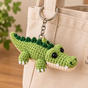 Puede incluir: Un llavero de cocodrilo tejido a ganchillo en tonos verde claro y oscuro con ojos de plástico negro y detalles blancos, unido a una anilla de metal plateado. Cuelga de un bolso de lona beige con un parche que dice Good Things Take Time.
