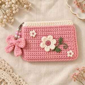 Puede incluir: Un monedero rectangular de ganchillo en color rosa con un borde superior crema. La parte frontal presenta aplicaciones florales blancas con centros rosas y hojas verdes. Un lazo de ganchillo rosa y un abalorio están unidos a una anilla.