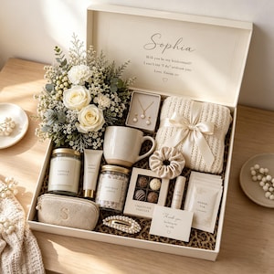 May include: A gift box containing a bouquet of white roses, a ceramic mug, a vanilla candle, bath salts, a jewelry set with a necklace and earrings, a knit blanket with a ribbon, a scrunchie, chocolates, lip balm, tea, a pouch with an S, and a hair clip.