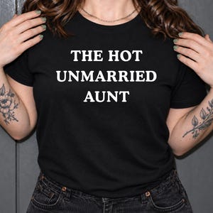 May include: A black short-sleeved t-shirt featuring the white text THE HOT UNMARRIED AUNT printed in a bold, sans-serif font across the chest. The garment has a classic crew neckline and is constructed from a solid black fabric material.