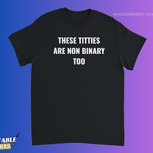 May include: A black short-sleeved t-shirt featuring the white text "THESE TITTIES ARE NON BINARY TOO" printed in a bold, sans-serif font across the center of the chest. The garment has a crew neckline and a standard, straight-cut silhouette.