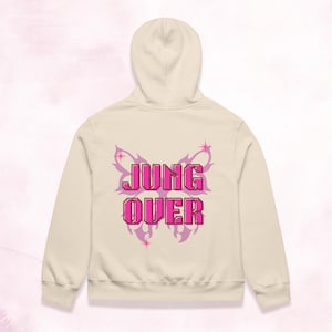 May include: A cream-coloured hooded sweatshirt featuring a large, light purple butterfly graphic on the back. Centered over the butterfly is the text JUNG OVER in a bold, pixelated, bright pink font, accented by small four-pointed star icons.