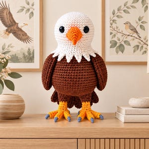Può includere: Un peluche a forma di aquila calva realizzato all'uncinetto con testa bianca, becco arancione brillante, ali e corpo marrone scuro e zampe gialle con artigli azzurri. Il peluche presenta occhi neri rotondi e una trama in filato.
