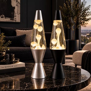 May include: Two lava lamps with conical bases and caps, one in a metallic silver finish and the other in a matte black finish. Both lamps feature clear glass vessels containing white wax blobs that are illuminated from within the base.