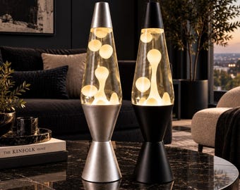 May include: Two lava lamps with conical bases and caps, one in a metallic silver finish and the other in a matte black finish. Both lamps feature clear glass vessels containing white wax blobs that are illuminated from within the base.