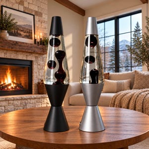 May include: Two lava lamps with conical bases and caps. The left lamp features a matte black finish with dark liquid and wax. The right lamp has a metallic silver finish with dark liquid and wax. Both lamps have clear glass bodies.