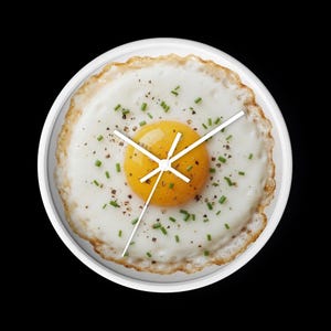 以下が含まれることがあります： 鮮やかな黄色の卵黄と白い卵白を持つ目玉焼きの高解像度プリントが施された丸い掛け時計。表面には緑色のチャイブと黒胡椒が散らされています。中央には3本の細い白い時計の針が取り付けられています。