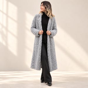 May include: A long, light gray coat featuring a textured, nubby knit fabric with long sleeves and an open front. The coat is worn over a black high-neck top and black trousers, creating a layered look with a consistent, neutral color palette.