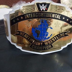 Puede incluir: Un cinturón de campeón de lucha libre WWE Intercontinental Heavyweight con una gran placa central de metal dorado, un gráfico de globo terráqueo azul, detalles en esmalte negro y correas de cuero blanco. El texto dice INTERCONTINENTAL HEAVYWEIGHT WRESTLING CHAMPION.
