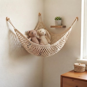 Puede incluir: Una hamaca de esquina de macramé color crema suspendida por tres ganchos de madera, que contiene un oso de peluche marrón y un conejo de peluche color crema. La hamaca presenta un diseño de red tejida y está montada en la esquina de una habitación.